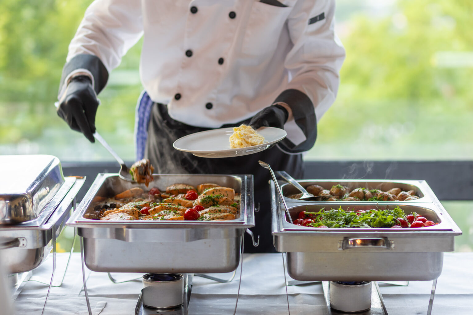 A chef in a white uniform carefully serves a plate of grilled chicken and fresh vegetables from a buffet setup during a catering event in a bright venue..